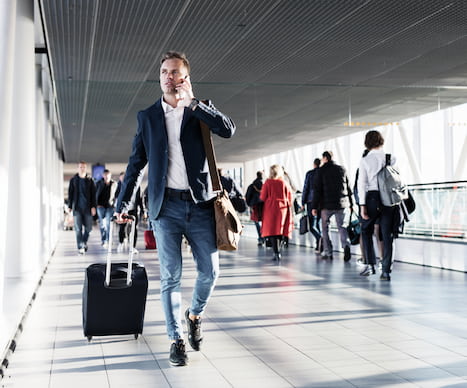 a businessman talks on the phone while pulling a suitcase through an airport hallway